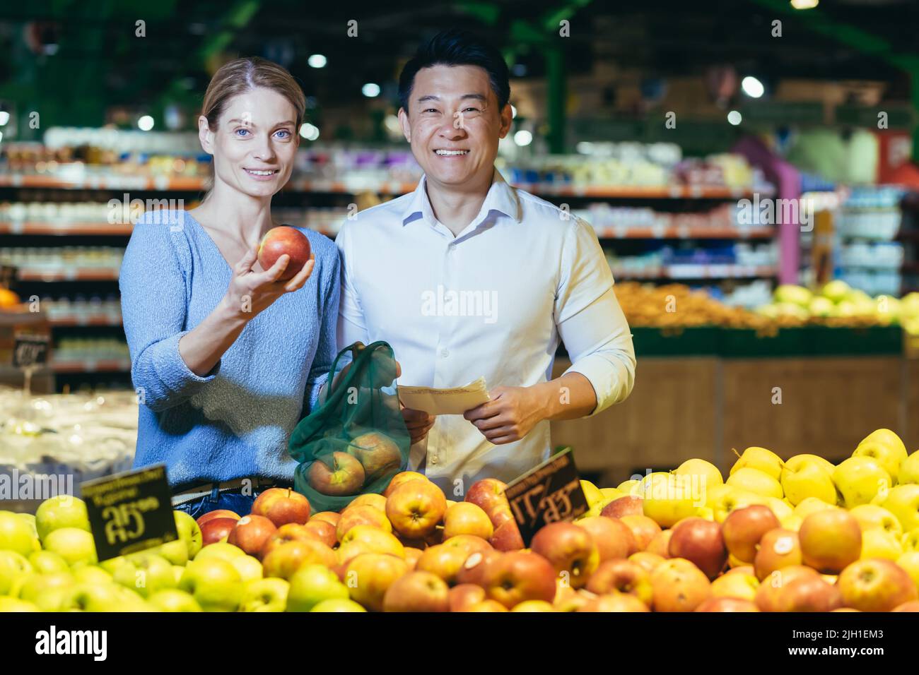 Porträt einer jungen multiethnischen Familie, eines asiatischen Mannes und einer asiatischen Frau beim Einkaufen im Supermarkt, lächelnd und vor die Kamera schauend, wählt das Paar Apfelfrüchte Stockfoto