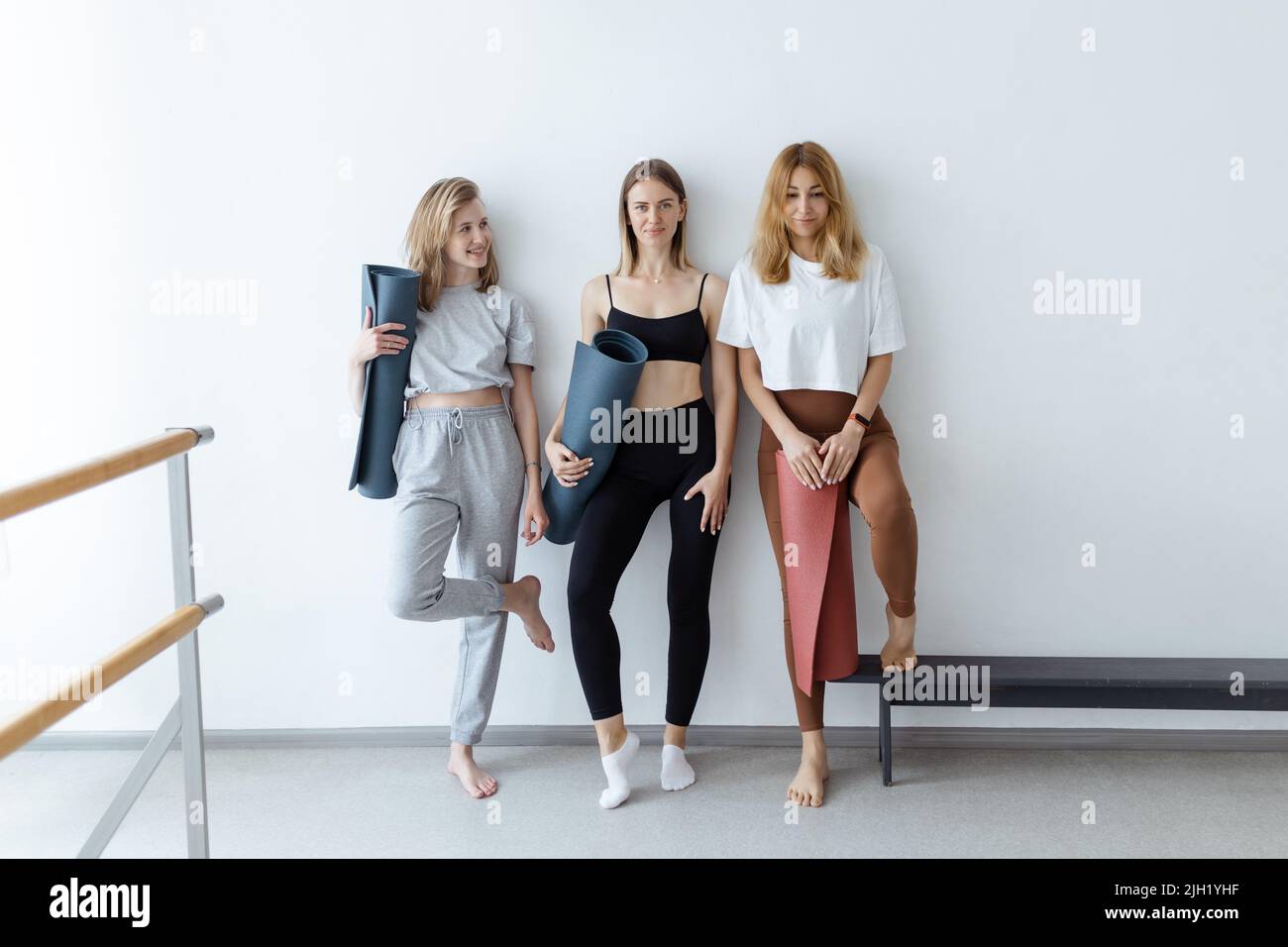 Eine Gruppe junger Sportmädchen mit Yogamatten, die an einer weißen Wand stehen. Freundinnen im Fitnessstudio, die sich nach dem Fitness- oder Yoga-Training drinnen entspannen Stockfoto