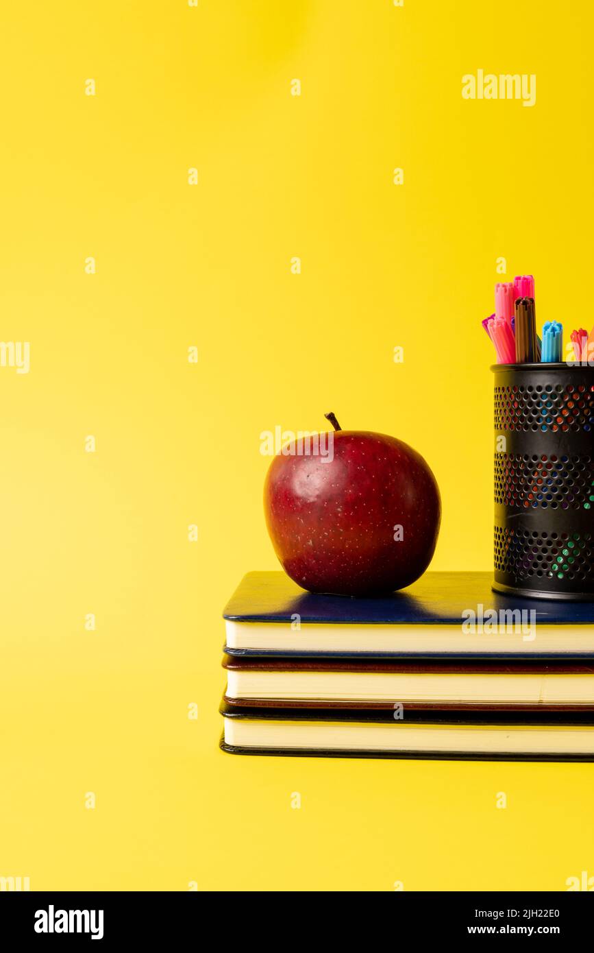Vertikale Zusammensetzung von Bücherstapel, Filzstiften im Behälter und Apfel auf gelbem Hintergrund Stockfoto