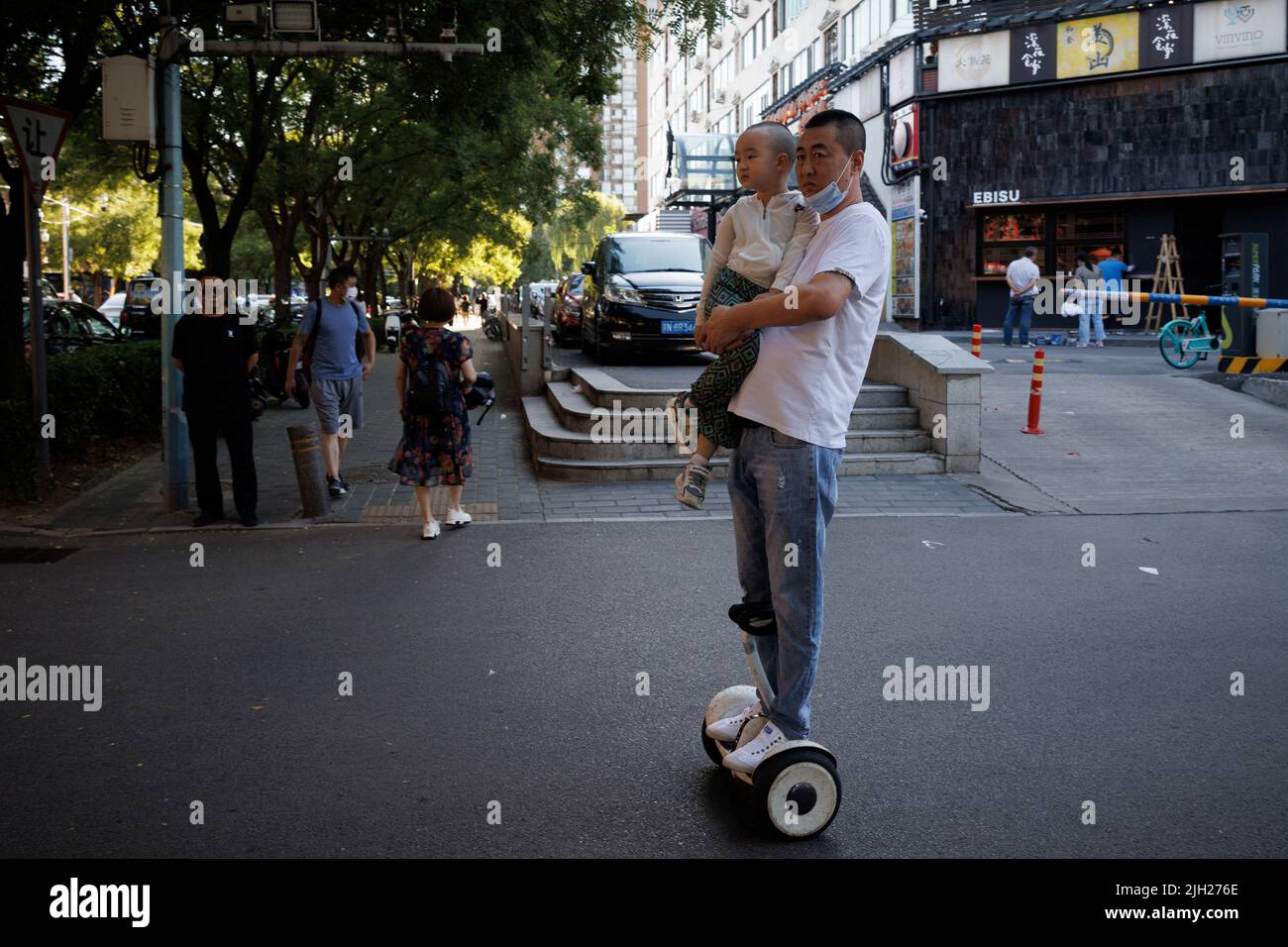 Ein Mann trägt ein Kind, während er auf einem Hoverboard in einer Wohneinkaufsstraße in Peking, China, fährt, 14. Juli 2022. REUTERS/Thomas Peter Stockfoto