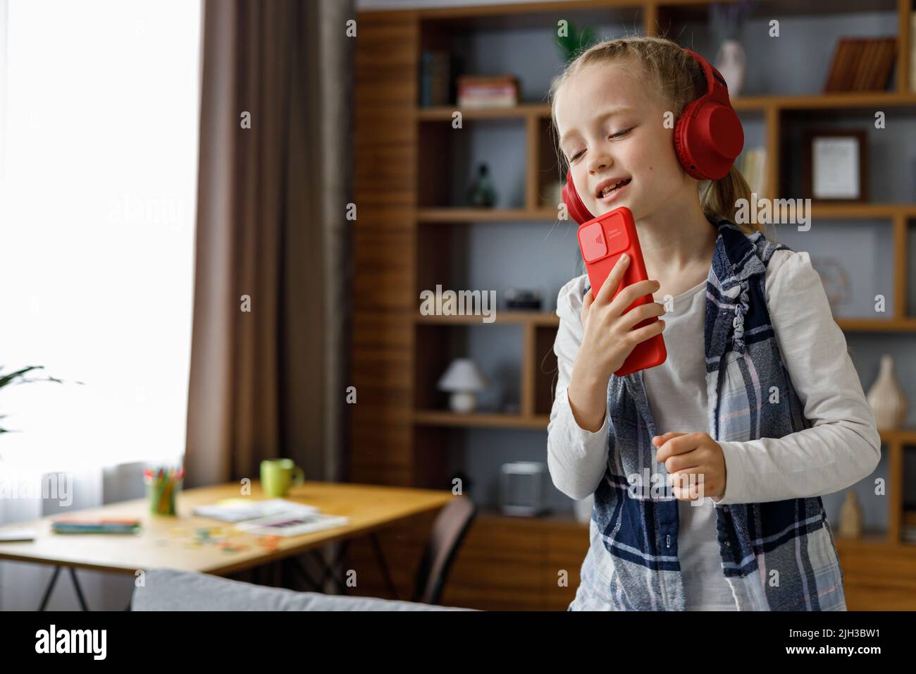 Glückliches kleines Mädchen mit roten Ohrhörern, das mit einem Smartphone wie einem Mikrofon tanzt und singt. Nettes Kind mit Spaß springen auf Trainer. Freizeit zu Hause Stockfoto
