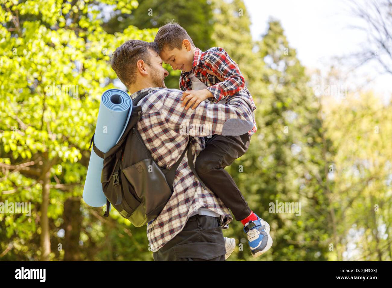 Fröhlicher kaukasischer Vater mit Rucksack, der den kleinen Sohn auf den Händen im Park am Sommertag hält. Stockfoto
