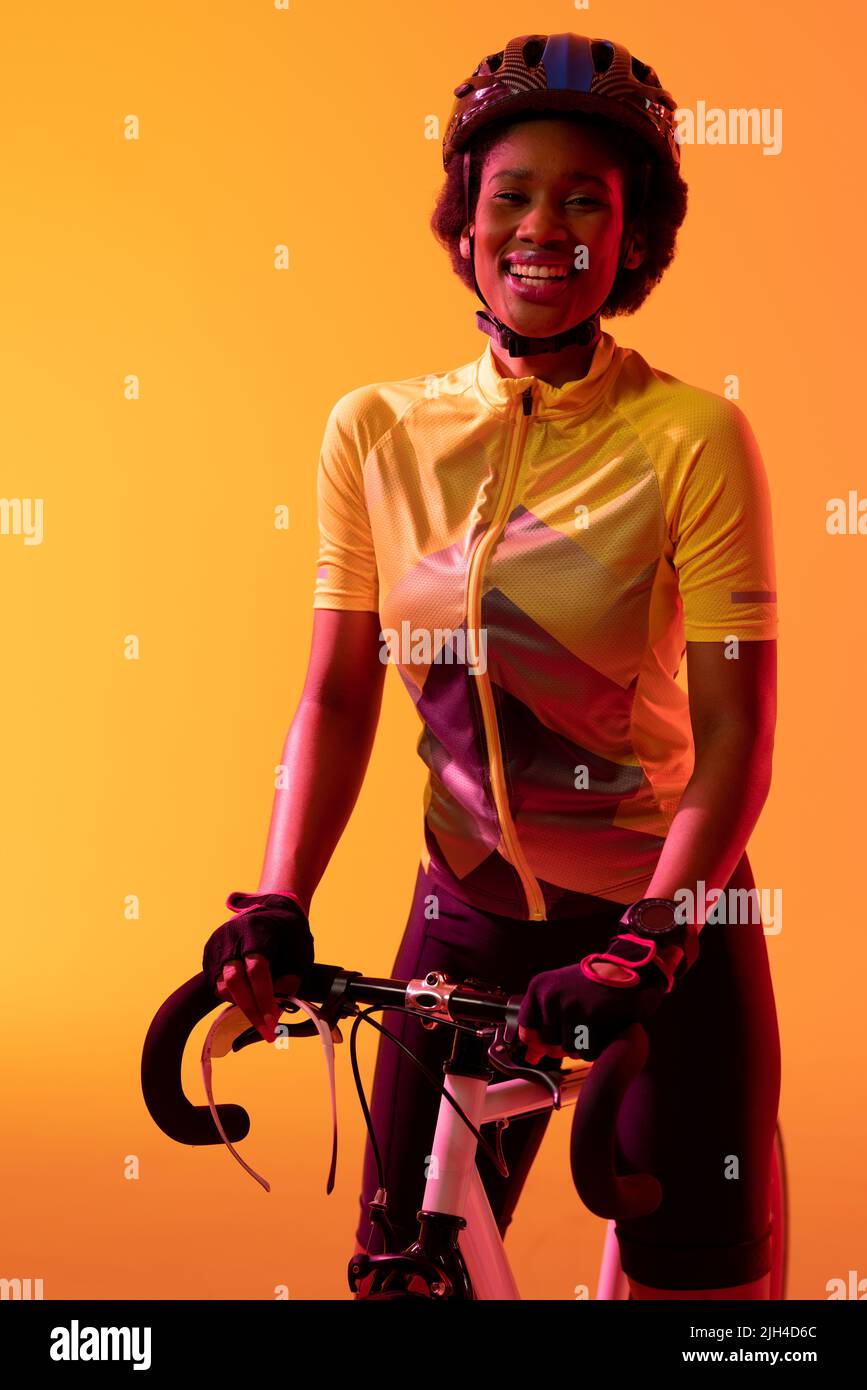 Vertikales Bild einer glücklichen afroamerikanischen Radfahrerin mit Fahrrad in neonoranger Beleuchtung Stockfoto
