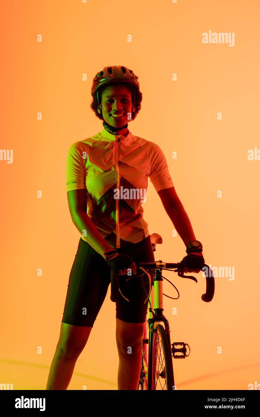 Vertikales Bild einer glücklichen afroamerikanischen Radfahrerin mit Fahrrad in neonoranger Beleuchtung Stockfoto