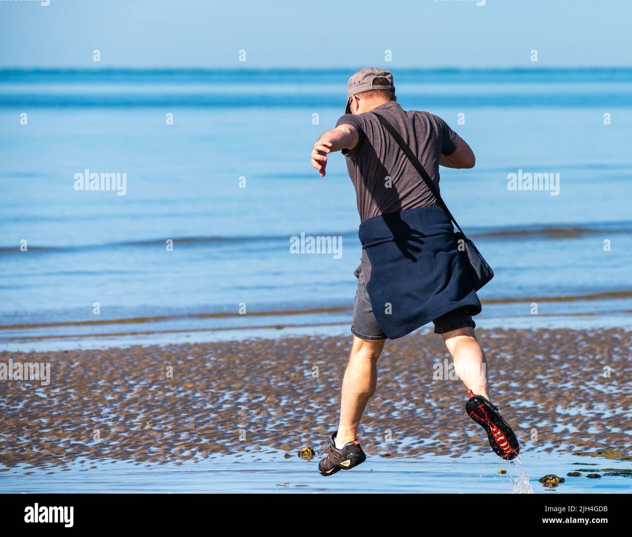 Littlehampton, West Sussex, Großbritannien. Freitag, 15.. Juli 2022. Ein Mann, der an einem weiteren sehr warmen und sonnigen Morgen an der Südküste über einen nassen Fleck am Strand springt. Kredit: Geoff Smith/Alamy Live Nachrichten Stockfoto