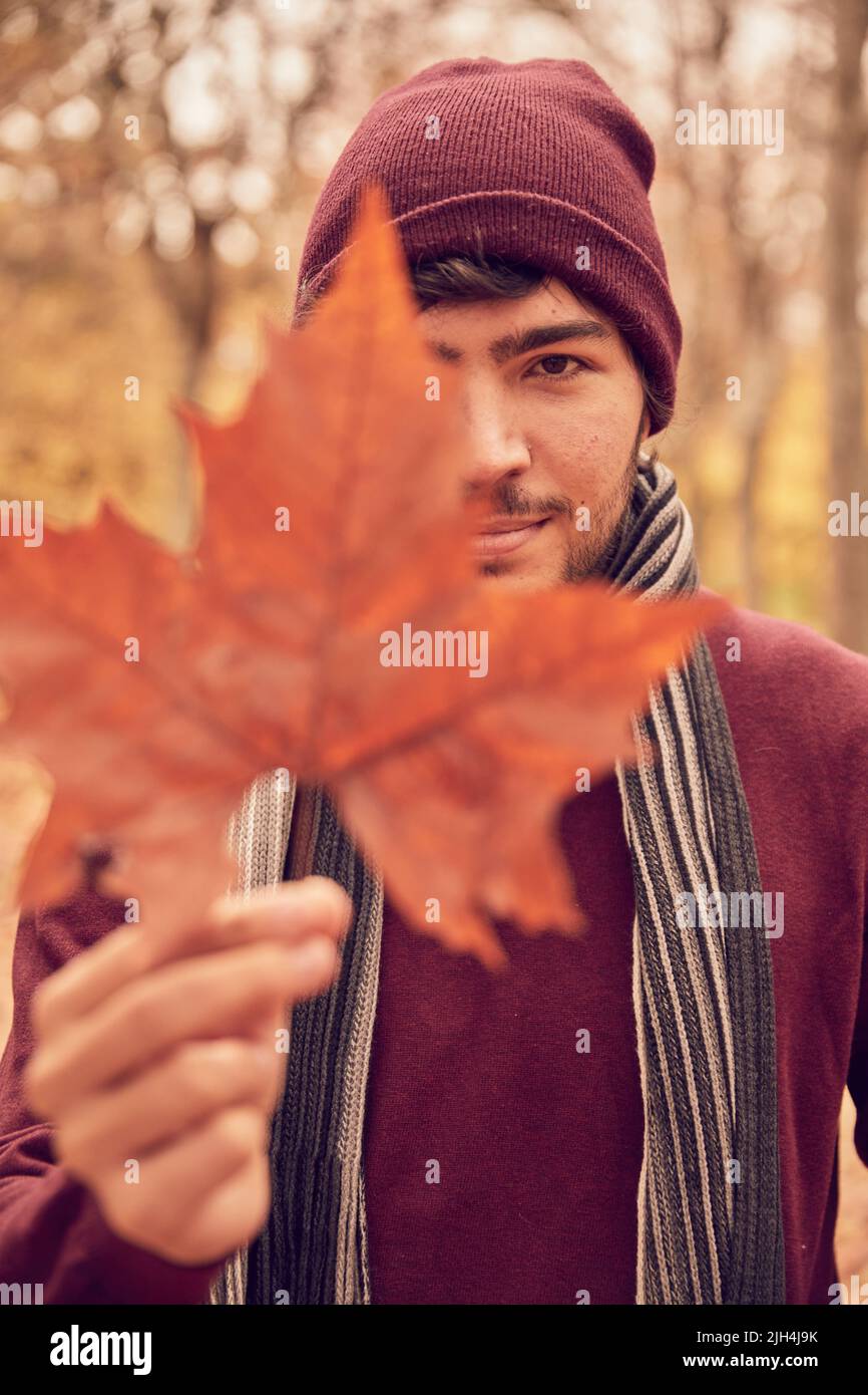 Weißer kaukasischer Mann in kastanienbraunen Mützchen und Pullover und grauem und schwarzem Schal, der die Kamera anschaut, die ein braunes fünfzackig Blatt mit einem Auge in einem Par umklammert Stockfoto