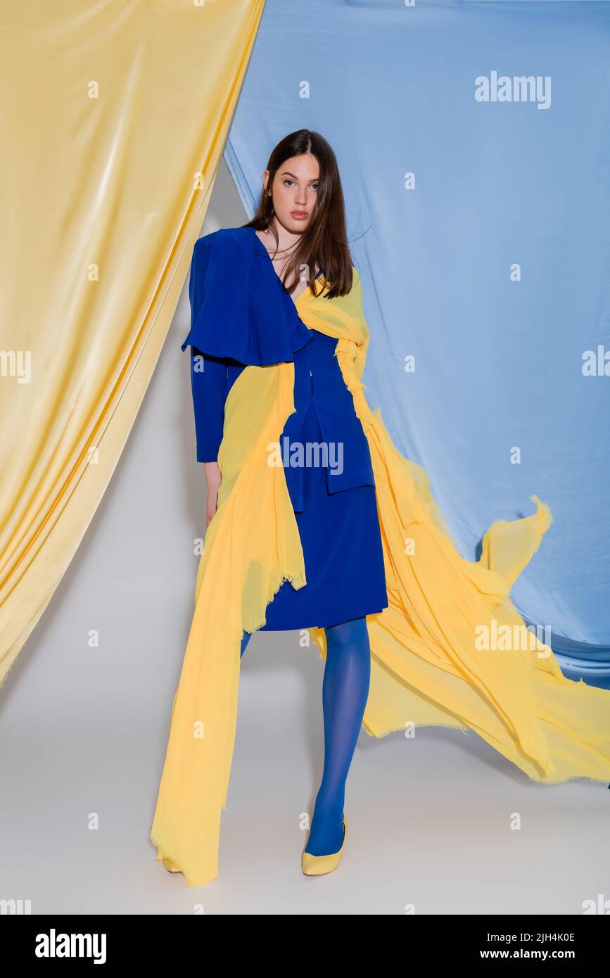 Volle Länge der jungen ukrainischen Frau in Farbe Block Kleid posiert in der Nähe von blauen und gelben Vorhängen Stockfoto