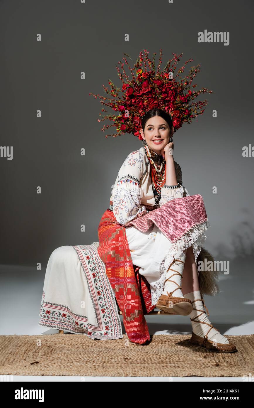 Volle Länge der glücklichen ukrainischen Frau in traditioneller Kleidung und roten Kranz mit Blumen und Beeren auf grau Stockfoto