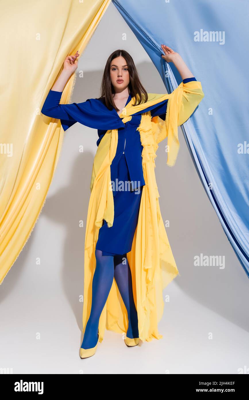 Volle Länge der hübschen ukrainischen Frau in Farbe Block Kleid posiert in der Nähe von blauen und gelben Vorhänge Stockfoto