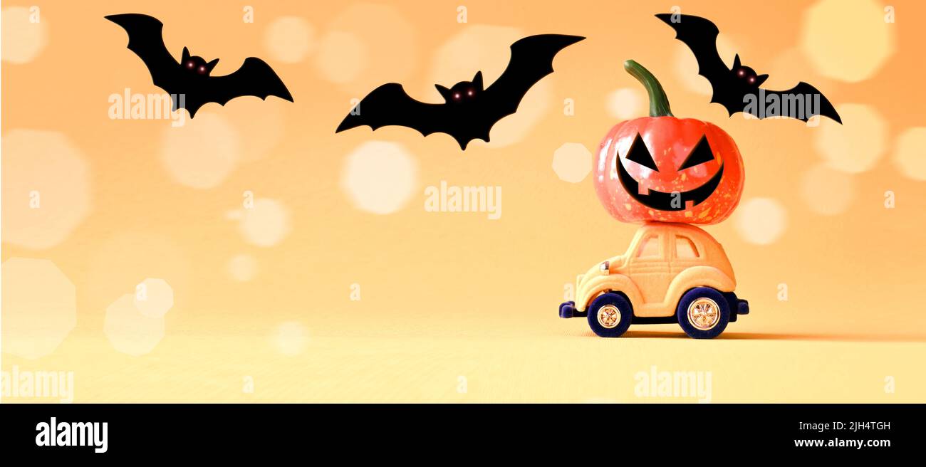 Spielzeugauto mit witzigem Kürbis auf dem Dach und Fledermäusen auf orangefarbenem Hintergrund. Platz für Text. Halloween Hintergrund. Stockfoto