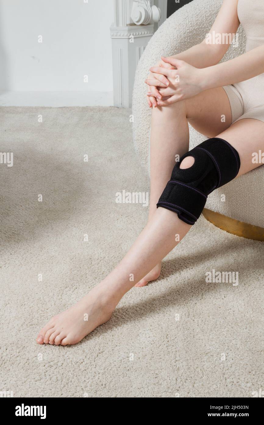 Schwarze Kniestütze auf einem Frauenbein. Mädchen in einer Orthese im Inneren des Hauses. Orthopädische anatomische Hosenträger zur Kniefixierung, Verletzungen und Stockfoto