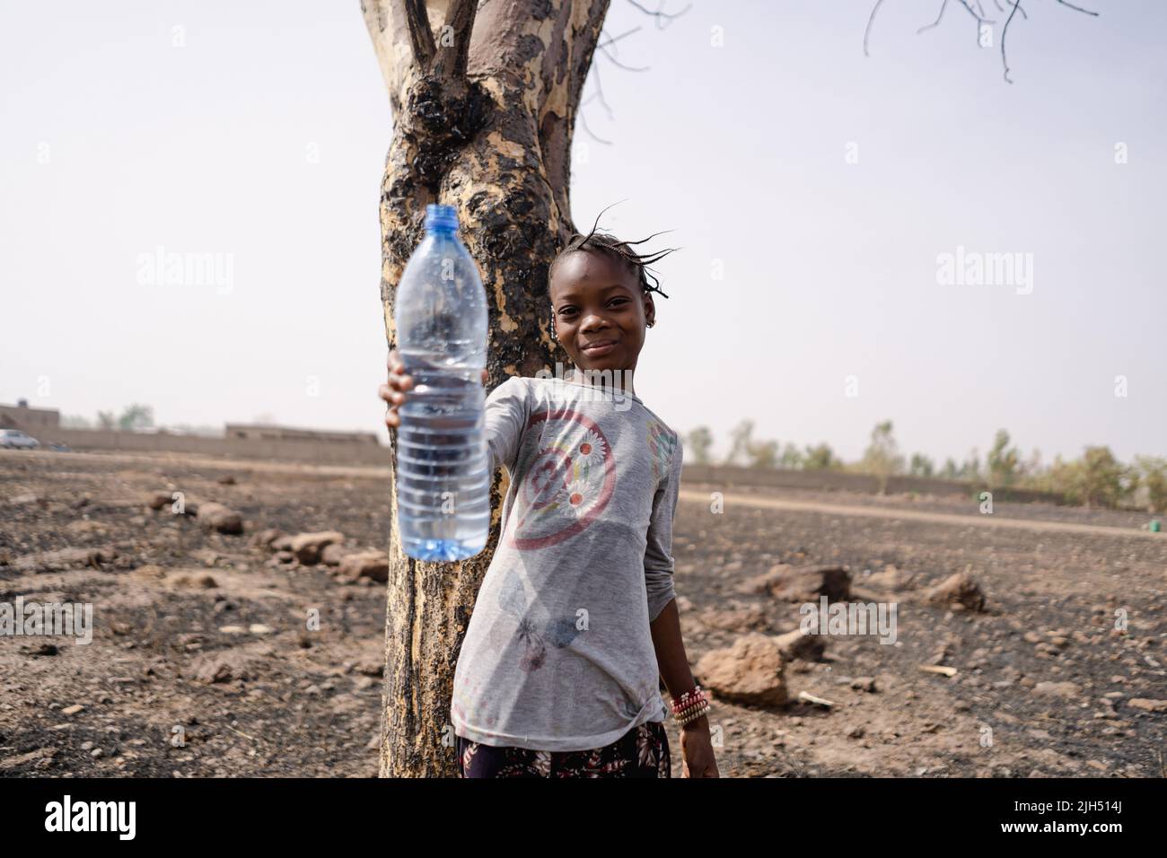 Schönes kleines afrikanisches Mädchen, das auf einem trockenen steinigen Feld steht und eine leere Plastikflasche hochhält, die den Wasserknappheit in der Region südlich der Sahara symbolisiert Stockfoto