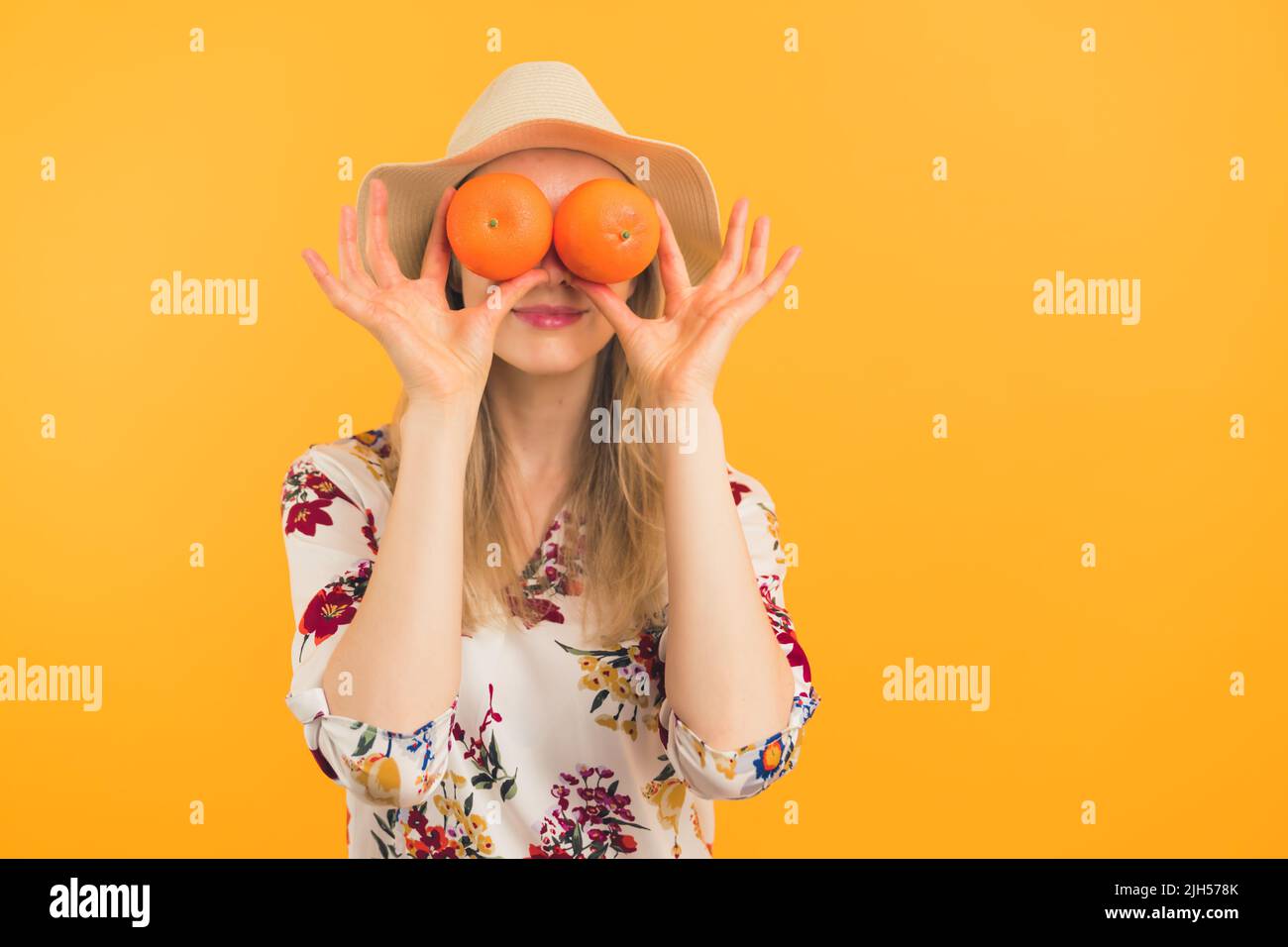 Gesunde Essgewohnheiten Konzept. Junge kaukasische langhaarige blonde Person in einem Hut und einer Blumenbluse, die die Augen mit zwei lebhaften Orangen bedeckt. Studio hat einen orangefarbenen Hintergrund aufgenommen. Hochwertige Fotos Stockfoto