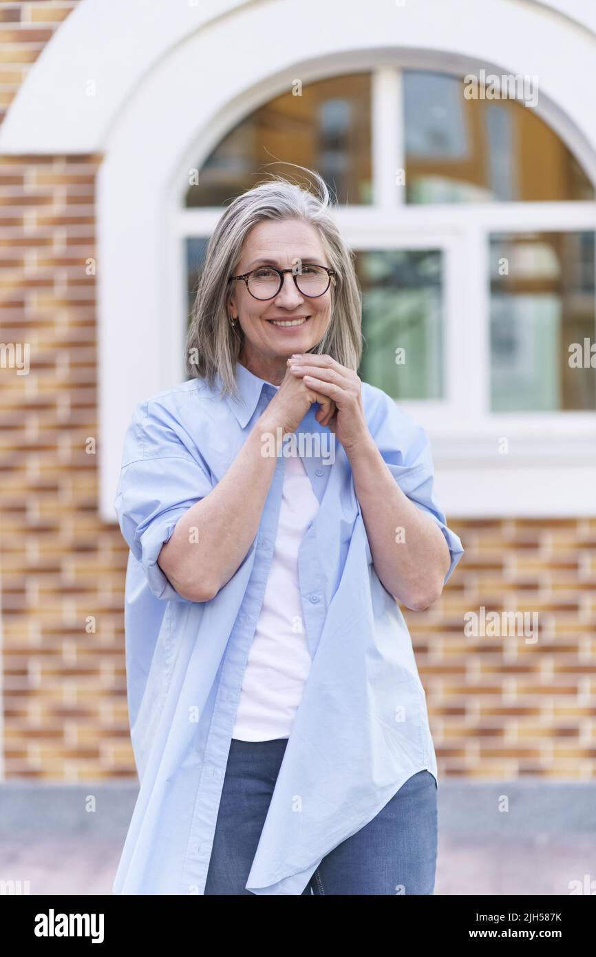 Porträt einer zarten Großmutter, die ihre Familie trifft. Reife graue Haare Frau trägt eine Brille im Freien, die im Ruhestand um die Welt reist. Reife Frau mit perfekter Haut trägt blaues Hemd. Stockfoto