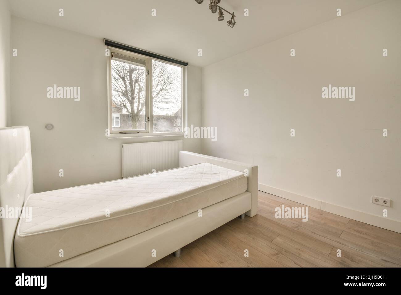 Komfortables Bett in einem kleinen, schmalen, minimalistischen Schlafzimmer mit weißen Wänden und Fenster in einem modernen Apartment Stockfoto