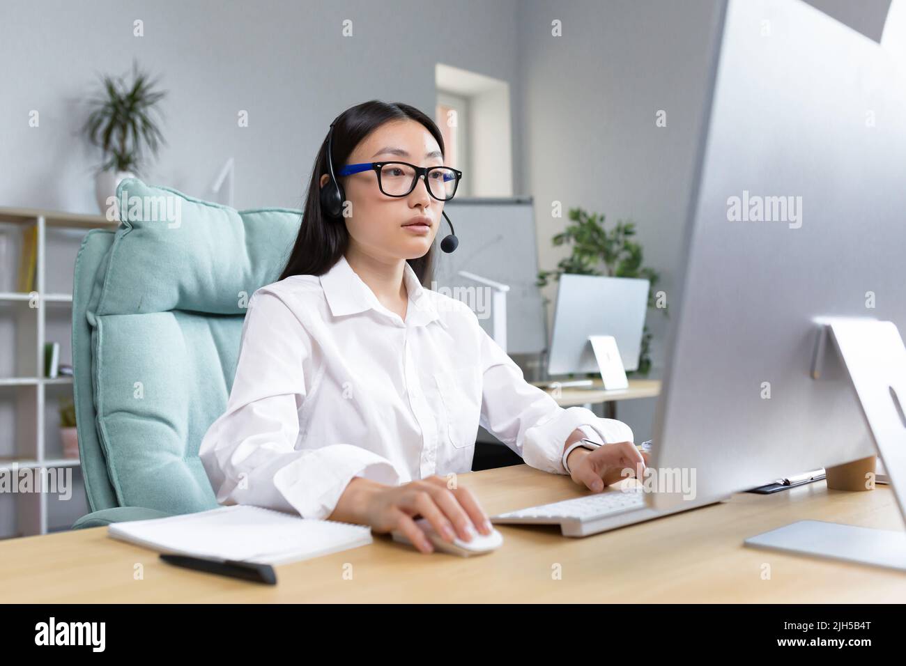 Online-Fernunterricht. Junge schöne asiatische Lehrerin mit Kopfhörern und Brille sitzt an einem Tisch an einem Computer und unterrichtet online eine Klassenstunde. Stockfoto