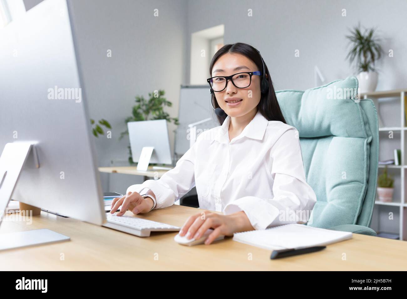 Online-Fernunterricht. Junge schöne asiatische Lehrerin mit Kopfhörern und Brille sitzt an einem Tisch an einem Computer und unterrichtet online eine Klassenstunde. Stockfoto