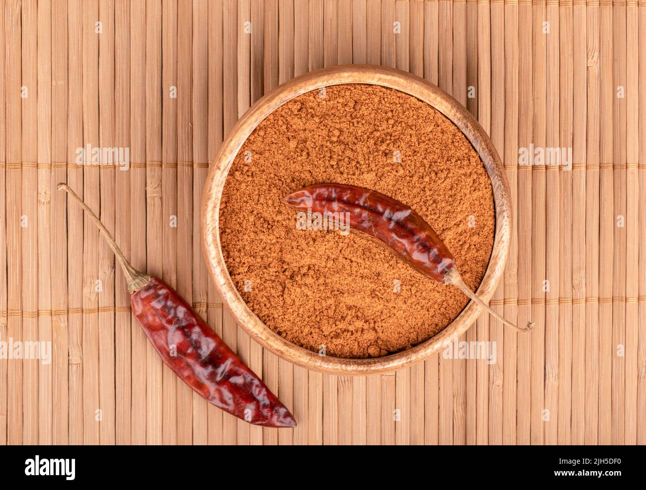 Draufsicht auf eine Holzschüssel mit gemahlenem rotem Pfeffer und Paprikaschoten auf einer Lebensmittelmatte. Eine Paprika-Schote liegt auf gemahlenem rotem Pfeffer. Stockfoto