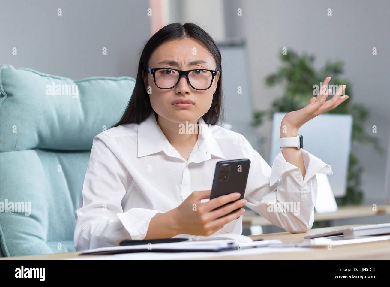 Besorgte junge, schöne asiatische Geschäftsfrau in Brille hält das Telefon in den Händen, schaut auf das Telefon, erhielt schlechte Nachrichten, breitet ihre Hände aus, überrascht, schockiert. Sitzen an einem Schreibtisch in einem modernen Büro. Stockfoto