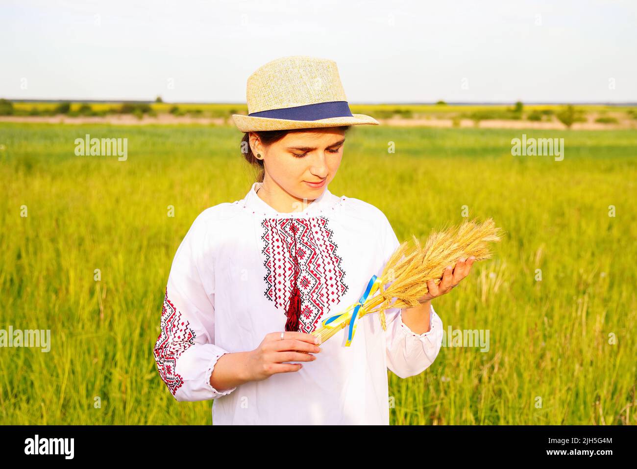 Unschärfe junge Frau in vyshywanka und Hut hält Bouquet von reifen goldenen Ähren gebunden auf der Wiese Natur Hintergrund. Flagge Ukraine Stockfoto