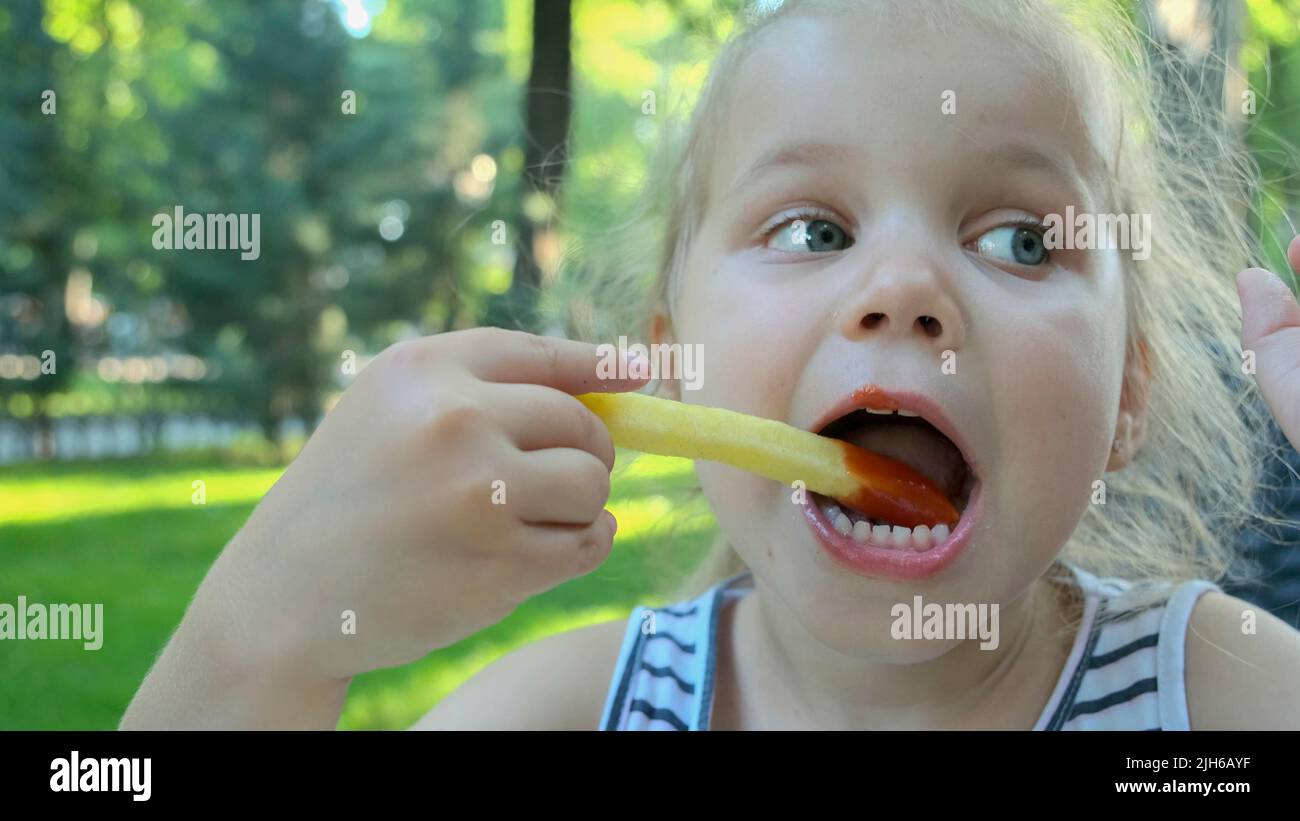 Kleine Mädchen essen pommes. Nahaufnahme des blonden Mädchens nimmt Kartoffelchips mit ihren Händen und probiert sie im Straßencafé im Park. Odessa Stockfoto