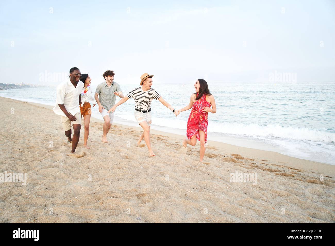 Fünf fröhliche Freunde, die Spaß am Strand haben, laufen im Sand zusammen, um einen glücklichen Wettbewerb zu führen. Junge Menschen genießen Sommerferien - Freundschaft Stockfoto