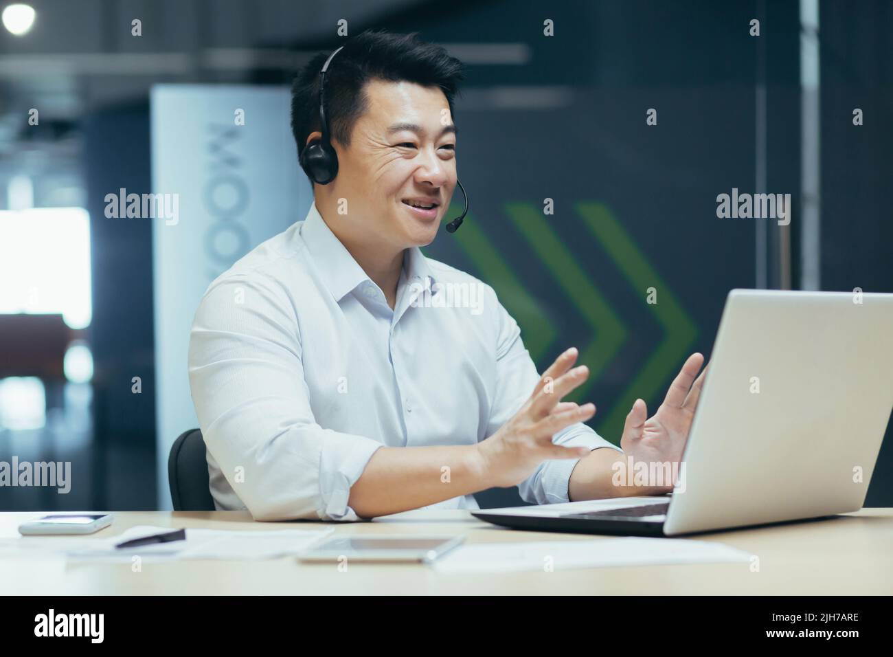 Ein junger, gutaussehender asiatischer Geschäftsmann mit Kopfhörern und Mikrofon sitzt im Büro an einem Laptop, führt eine Online-Konferenz durch, ein Remote-Meeting mit Kunden und Partnern. Erzählt, erklärt, lächelt Stockfoto