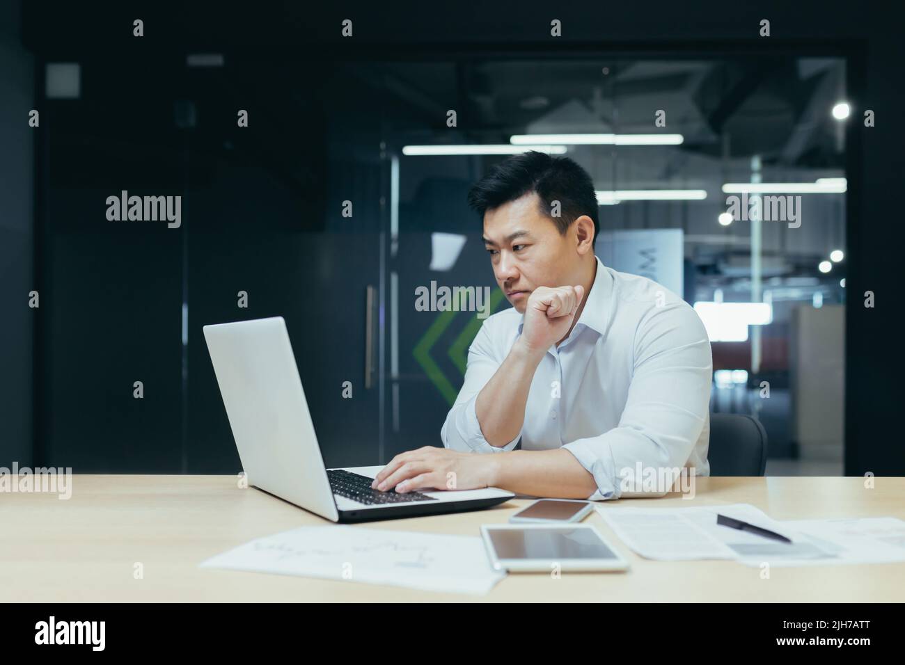 Ein junger, gutaussehender Mann, ein asiatischer Geschäftsmann, arbeitet mit einem Laptop und tippt. Sitzen an einem Schreibtisch in einem modernen Büro Stockfoto