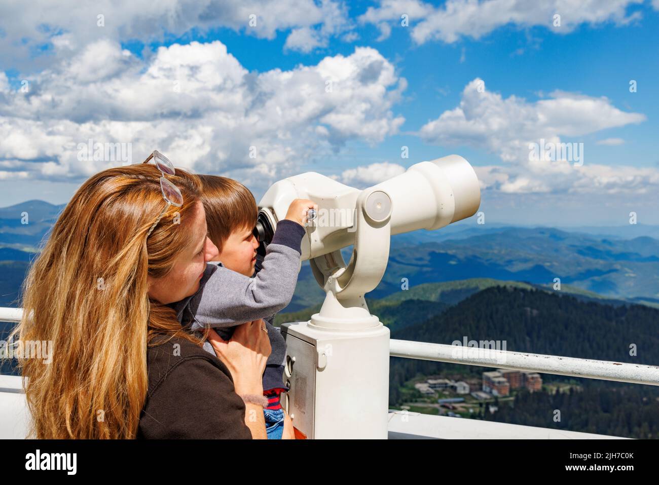 Eine fürsorgliche Mutter zeigt ihrem neugierigen kleinen Sohn die Landschaften im bewaldeten Tal der Rhodopen und den wolkigen Himmel durch ein Teleskop, auf einem Stockfoto