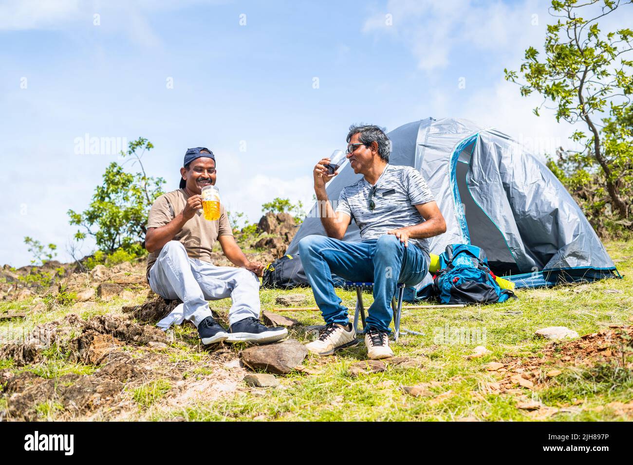 Glücklich lächelnde Wanderer, die sich beim Biertrinken gut unterhalten, während sie sich vor dem Zelt am Hügel unterhalten - Konzept der Freundschaft, Urlaub Stockfoto