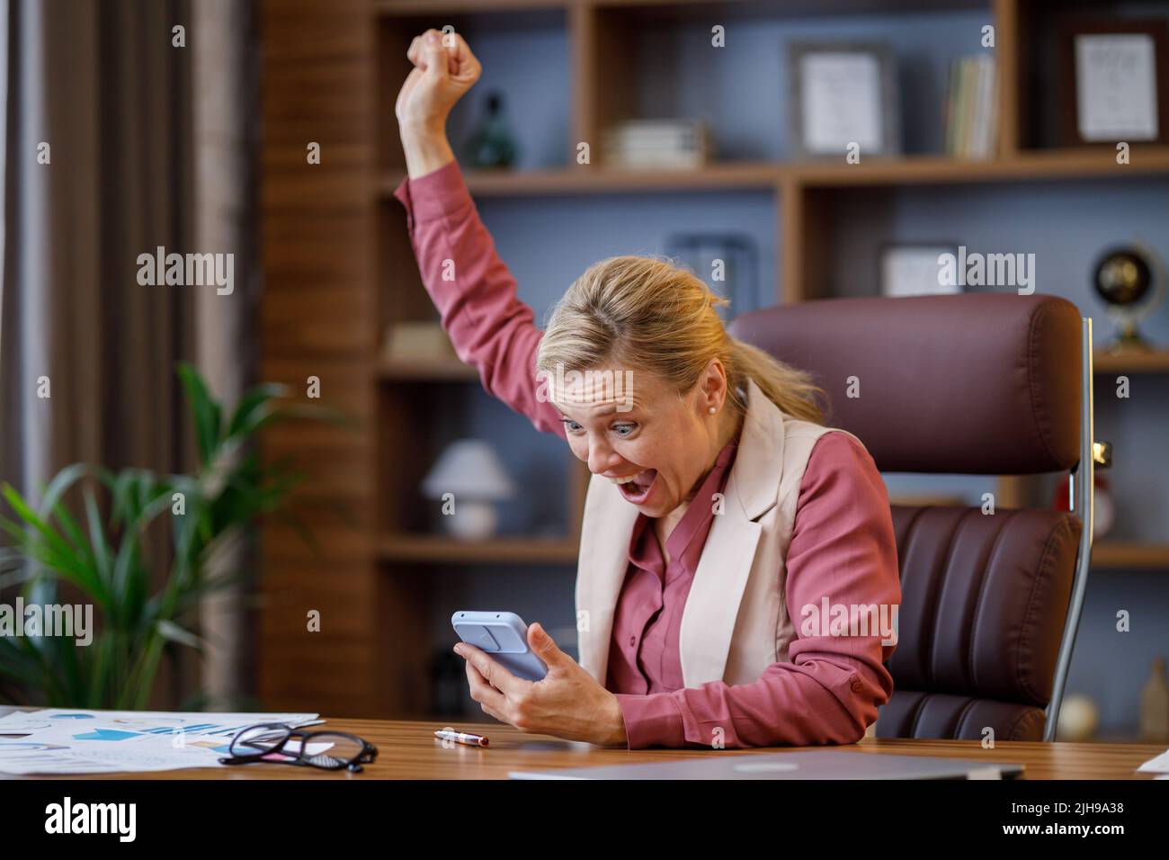 Glückliche Geschäftsfrau überrascht von den guten Nachrichten und macht ja Geste beim Blick auf Smartphone im Büro sitzen. Der Manager genießt den Erfolg Stockfoto