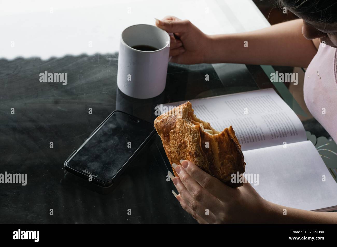 Schöne Latina Frau hält in ihrer linken Hand ein Sandwich, das sie isst, bevor sie ins Büro geht. Mädchen, das ein Buch liest, während sie Kaffee trinkt Stockfoto