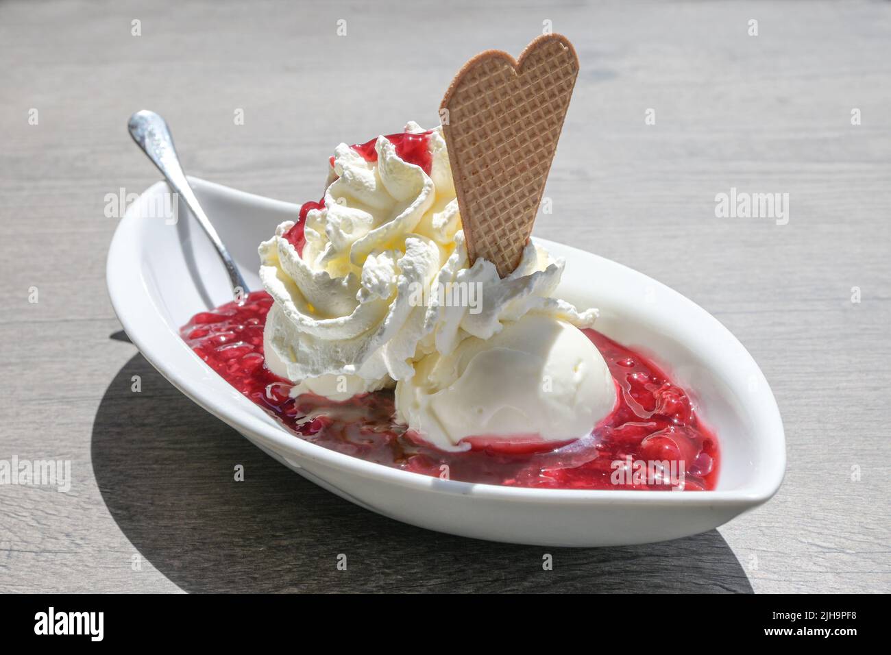 Eis mit Schlagsahne, Waffelplätzchen und rotem Fruchtgelee, in Deutschland rote Gruetze genannt, serviert in einer weißen Schüssel auf einem grauen Tisch auf einem sonnigen Summ Stockfoto