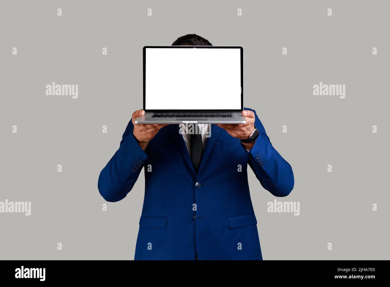Anonymer unbekannter Mann, der sein Gesicht hinter dem Laptop versteckt, mit weißem, leerem Display mit Platz für Werbung, in einem Anzug im offiziellen Stil. Innenaufnahme des Studios isoliert auf grauem Hintergrund. Stockfoto