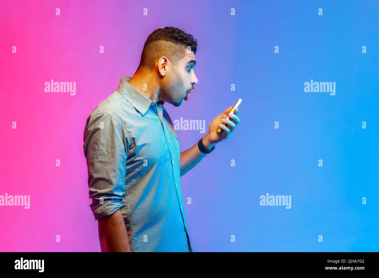Seitenansicht eines erstaunt überraschten Mannes im Hemd, der den Online-Service auf dem Mobiltelefon nutzt und Erstaunen und Schock zum Ausdruck bringt. Innenaufnahmen im Studio, isoliert auf farbigem Neonlicht-Hintergrund. Stockfoto