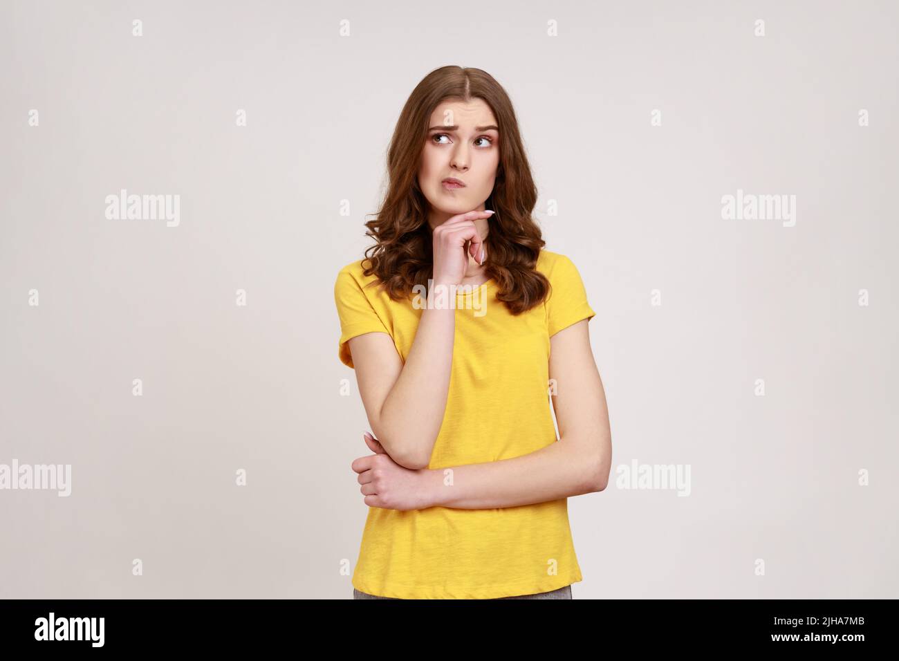 Nachdenklich schöne Teenager-Mädchen in lässiger Kleidung Zweifel, nicht sicher, mit unsicher verwirrt Gesicht, hält Kinn, schaut weg. Innenaufnahme des Studios isoliert auf grauem Hintergrund. Stockfoto