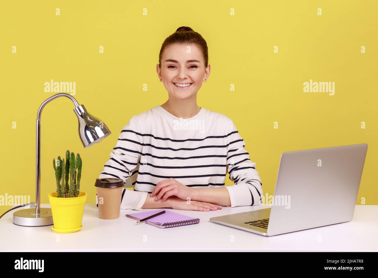 Glückliche, erfolgreiche Geschäftsfrau, die am Büroarbeitsplatz sitzt, lächelnd auf die Kamera schaut, zufrieden mit dem Job ist, optimistisch sieht. Studio-Innenaufnahme isoliert auf gelbem Hintergrund. Stockfoto