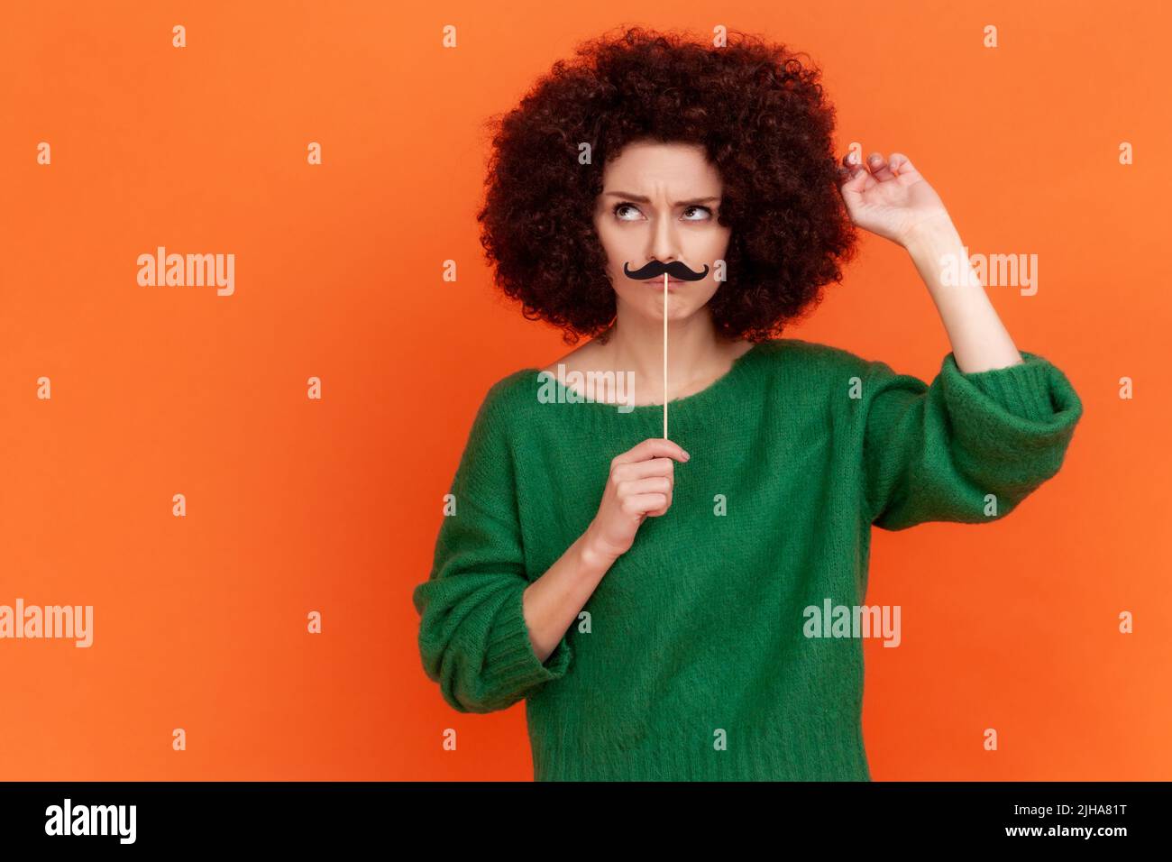 Nachdenkliche Frau mit Afro-Frisur trägt einen grünen Pullover im lässigen Stil mit festlicher Stimmung, steht mit Schnurrbart und ist tief in Gedanken. Innenaufnahme des Studios isoliert auf orangefarbenem Hintergrund. Stockfoto