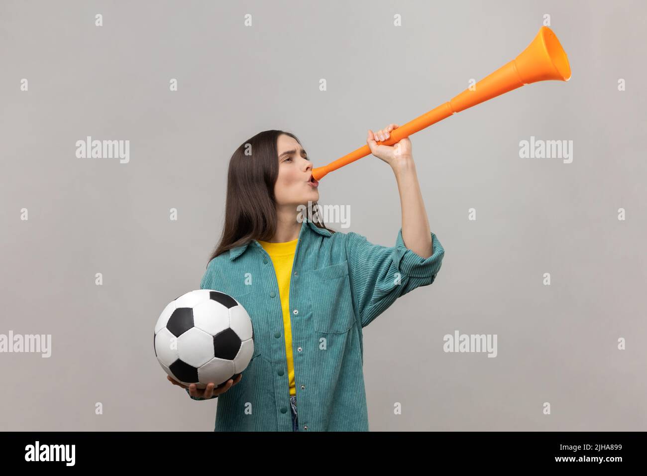 Seitenansicht einer aufgeregten Frau, die in Horn bläst und den schwarz-weißen Fußballball hält, um den Sieg der Lieblings-Fußballmannschaft zu feiern, in einer legeren Jacke. Innenaufnahme des Studios isoliert auf grauem Hintergrund. Stockfoto