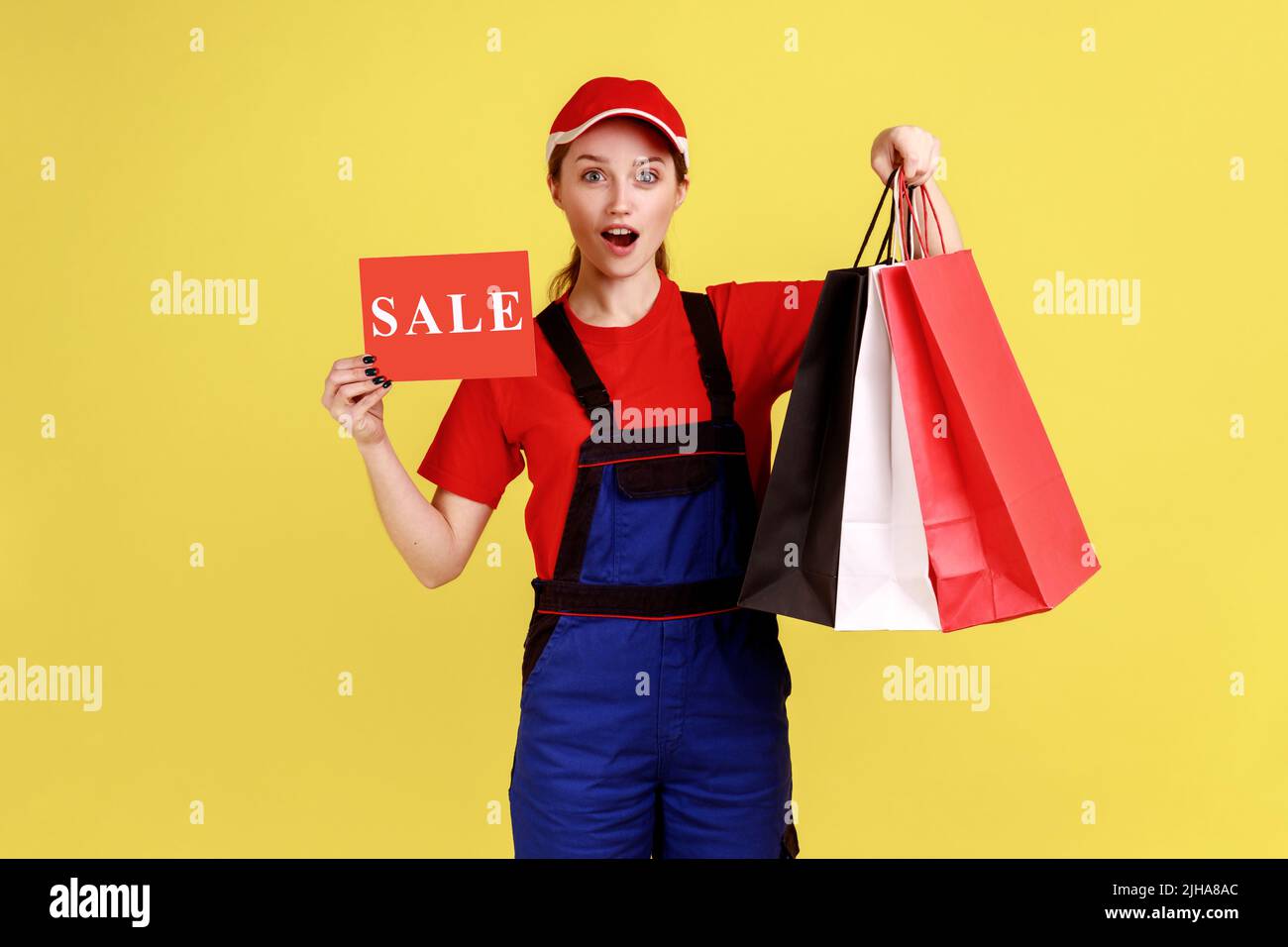 Portrait einer überraschten Lieferfrau, die mit Einkaufstaschen und Karte mit Verkaufsaufschrift steht, Overalls und roter Mütze trägt. Innenaufnahme des Studios isoliert auf gelbem Hintergrund. Stockfoto