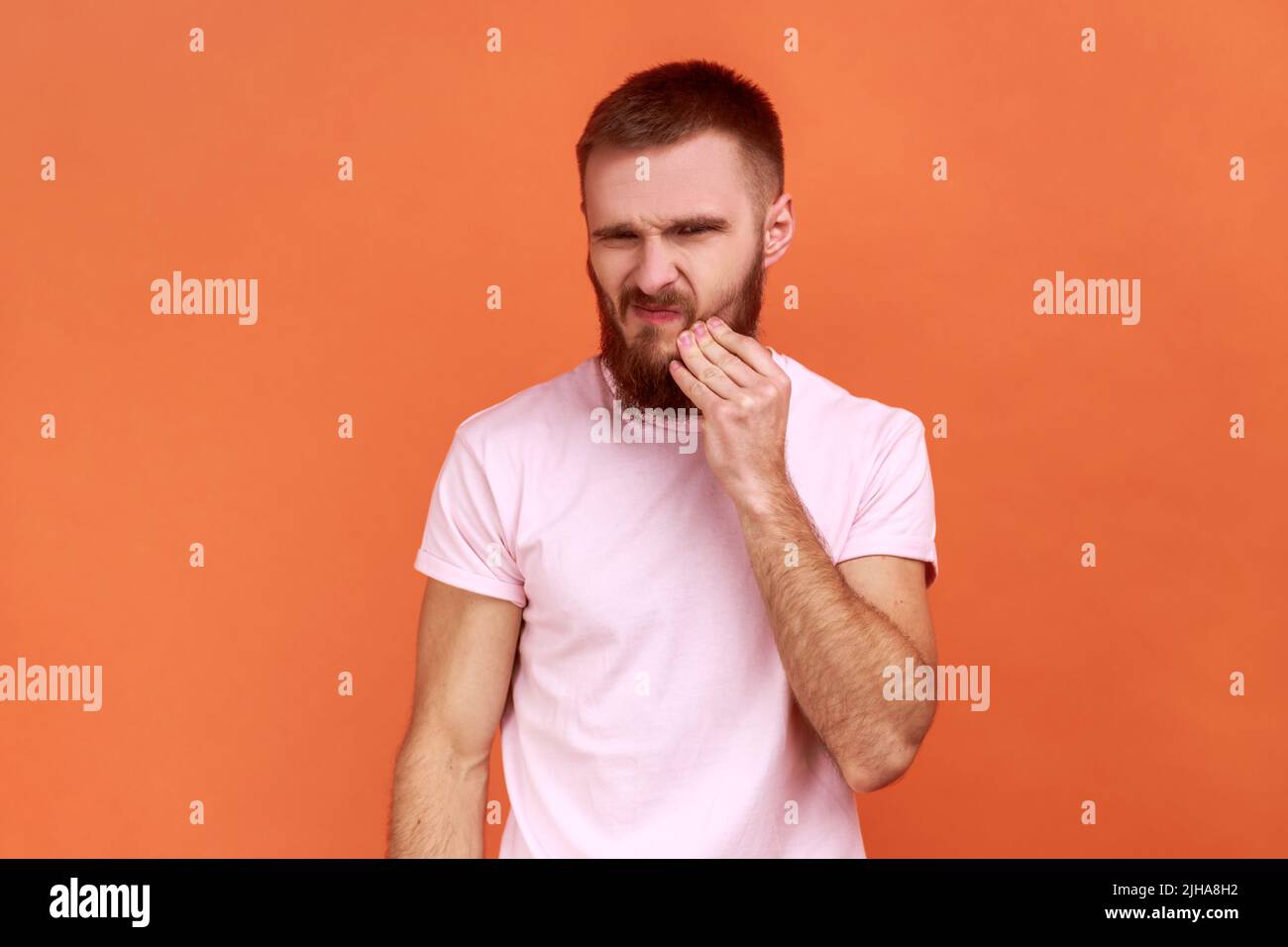 Zahnproblem. Porträt eines bärtigen Mannes, der seine Wange berührt und sich von schrecklichen Zahnschmerzen, empfindlichen Zähnen und einem rosa T-Shirt grimazt. Innenaufnahme des Studios isoliert auf orangefarbenem Hintergrund. Stockfoto