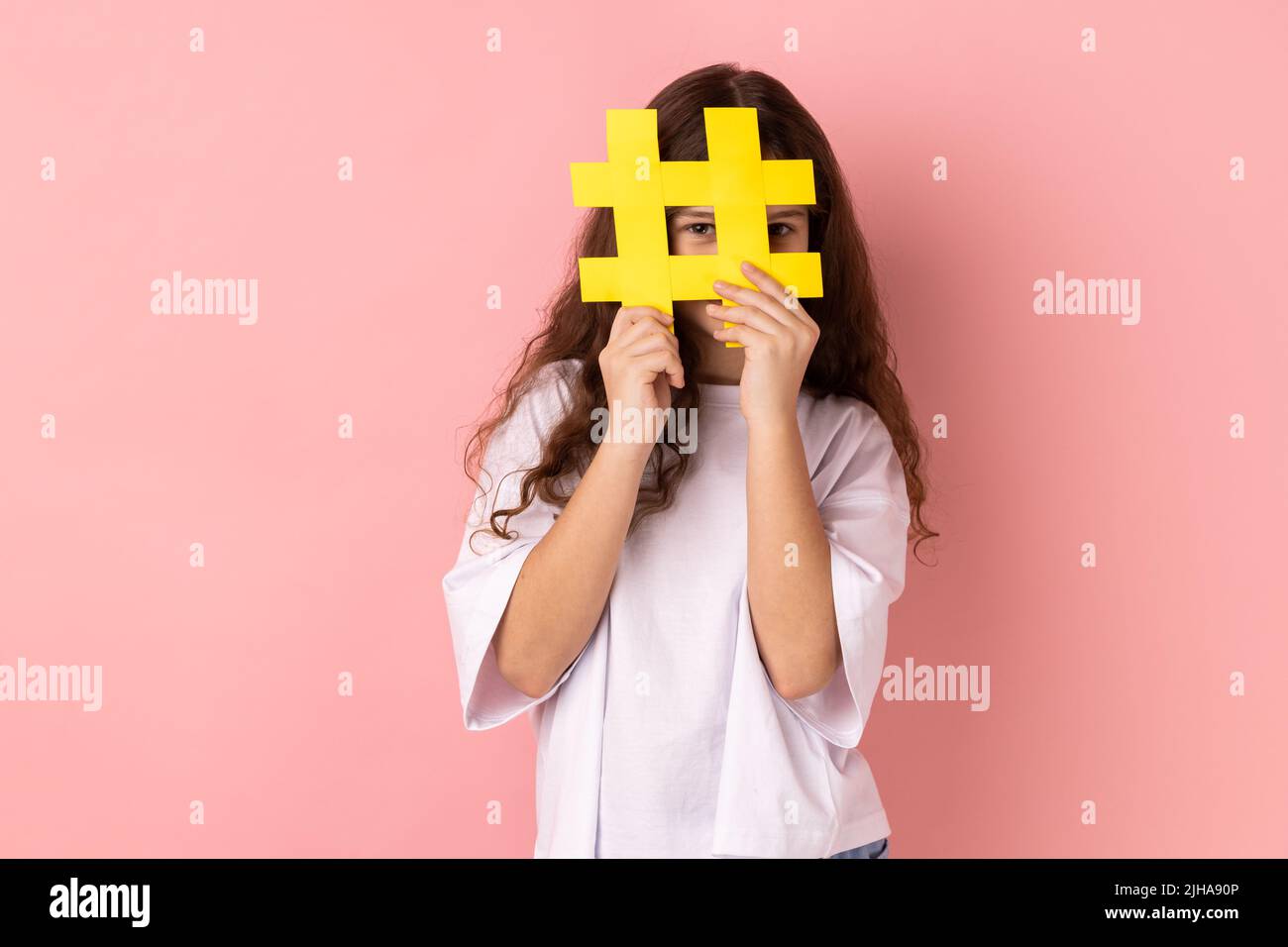 Kleines Mädchen trägt weißes T-Shirt bedeckt Gesicht mit Social-Media-Hashtag-Symbol, empfehlen, trendige Inhalte, beliebte Business-Blog folgen. Innenaufnahme des Studios isoliert auf rosa Hintergrund. Stockfoto