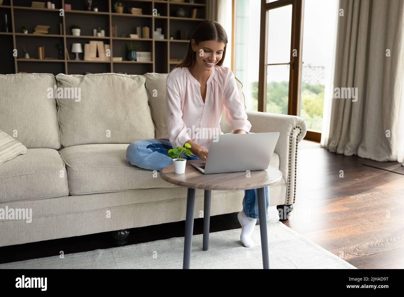 Eine lächelnde Frau sitzt mit einem Laptop auf der Couch am Tisch Stockfoto