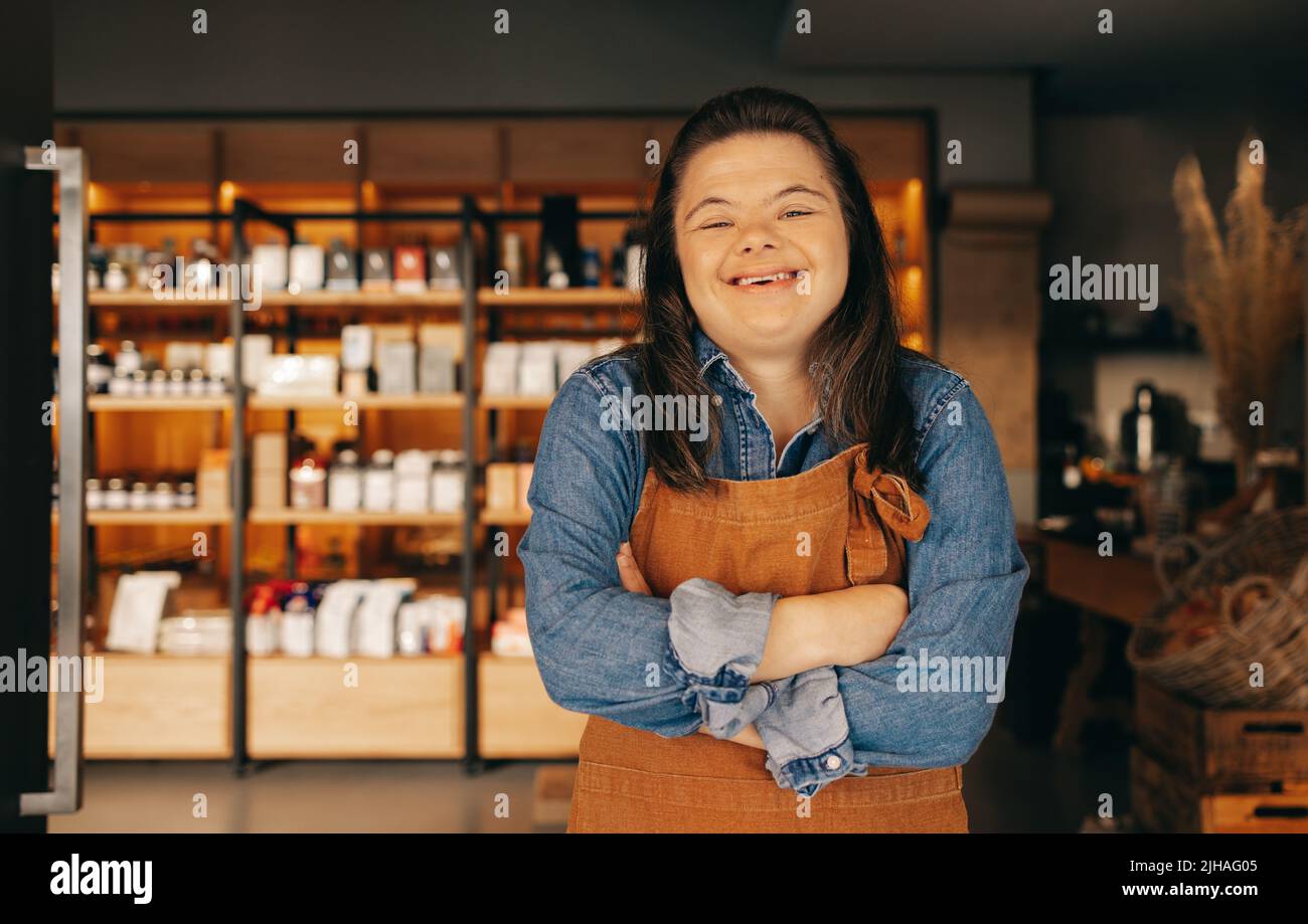Glückliche Frau mit Down-Syndrom lächelt, während sie am Eingang eines Delikatessenessenläden steht. Eine Frau mit einer intellektuellen Behinderung, die als Ladeninhaberin arbeitet Stockfoto