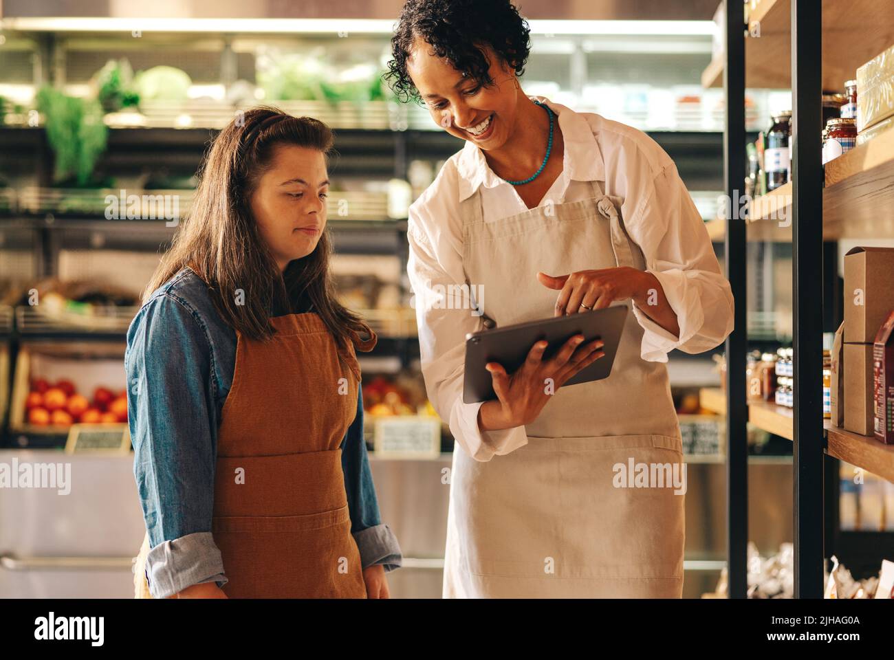 Glückliche Lebensmittelmanagerin, die ein digitales Tablet verwendet, während sie mit ihrem Mitarbeiter ein Gespräch führt. Weibliche Ladenbesitzerin, die eine Frau mit Down-Syndrom tr Stockfoto
