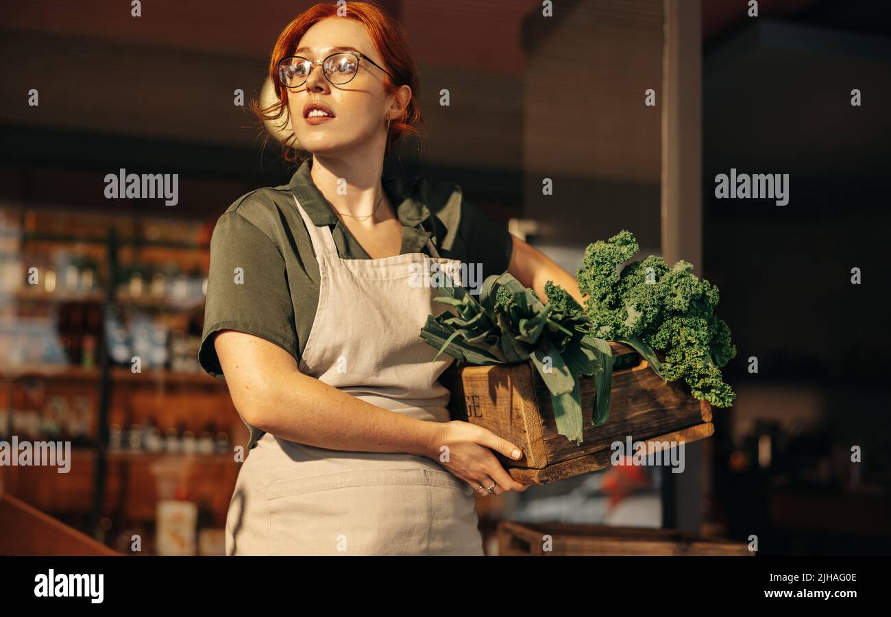 Weibliche Lebensmittelgeschäftbesitzerin schaut nachdenklich weg, während sie eine Schachtel mit frischem Bio-Gemüse in ihrem Laden hält. Junge Unternehmerin, die eine Stockfoto