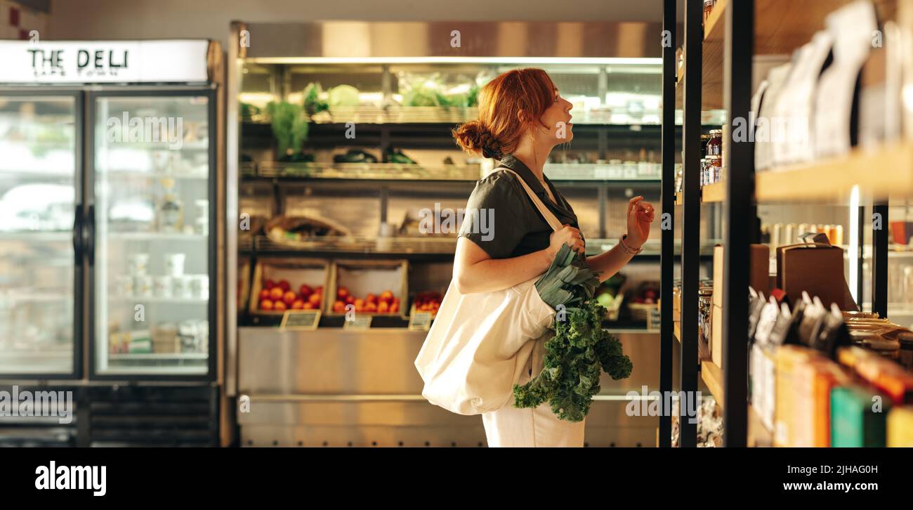 Weibliche Shopper, die Lebensmittel aus einem Regal auswählen, während sie eine Tasche mit Gemüse in einem Lebensmittelgeschäft tragen. Junge Frau macht ein paar Lebensmitteleinkäufe in Stockfoto