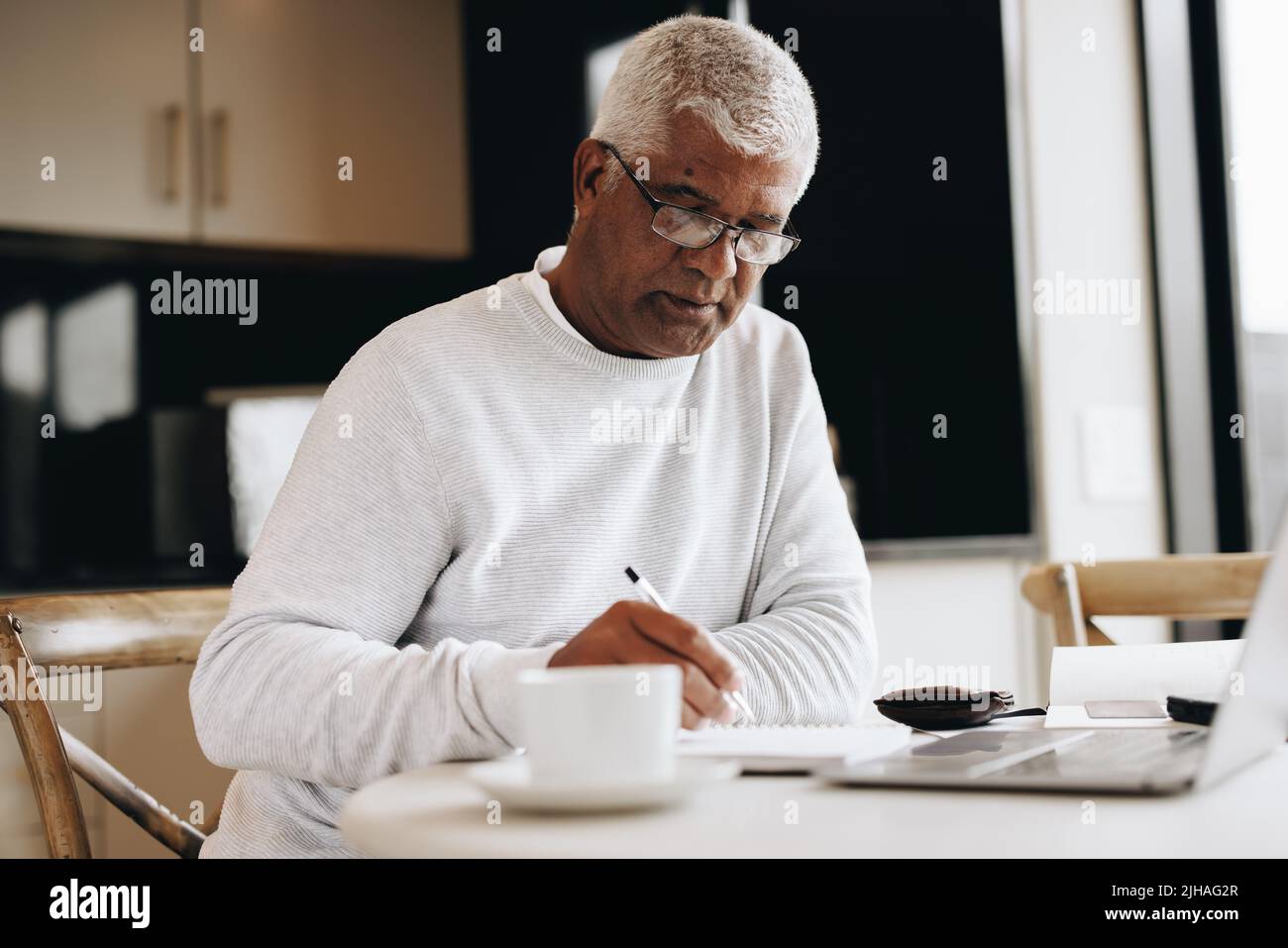 Ein leitender Geschäftsmann schreibt in einem Notizbuch, während er an einem Schreibtisch in seinem Haus sitzt. Reifer Geschäftsmann, der in seinem Tagebuch Pläne schmiedet, während er Remote Freela macht Stockfoto