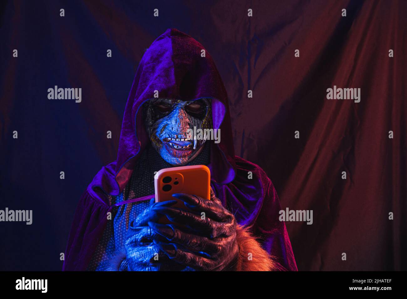 Portrait eines Zombies, der mit seinem Handy in einem Hemd und einem Kapuzenumhang gekleidet ist. Die Szene ist dunkel, beleuchtet von blauen und orangen Lichtern, das Gesicht des Monsters ist es Stockfoto