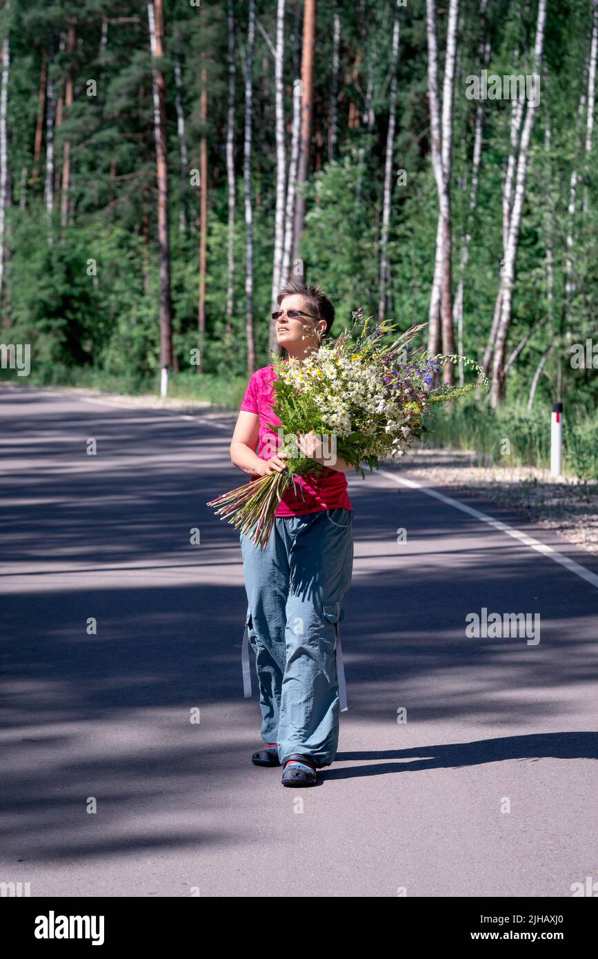 Ganzkörperportrait einer glücklichen lächelnden Frau mit einem riesigen Strauß wilder Blumen, die auf einer Waldstraße spazieren Stockfoto
